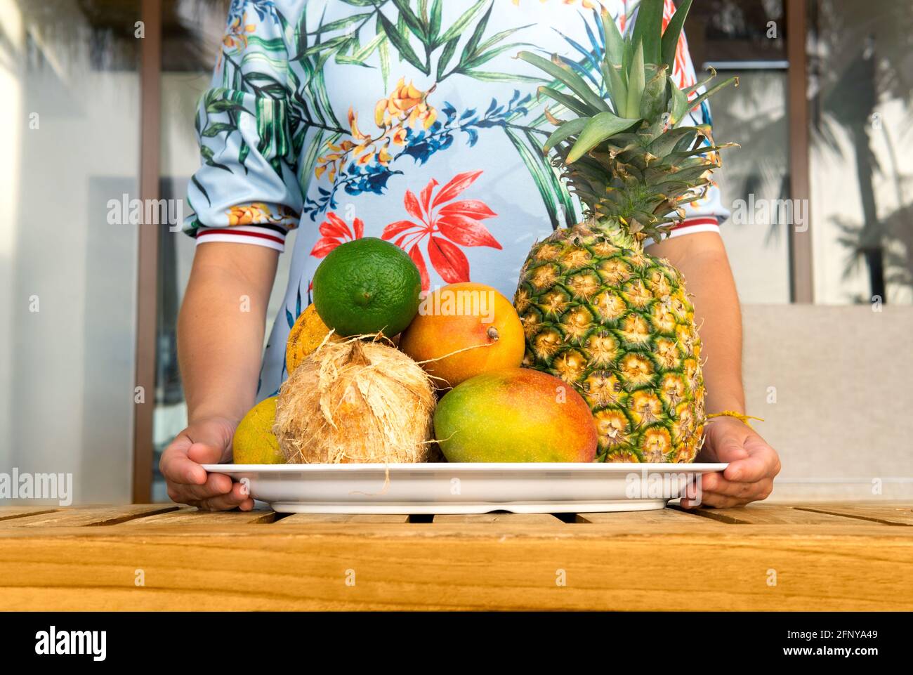 SANTA TERESA, COSTA RICA - 2. APRIL 2021: Weibchen in einem bunten Kleid, das ein großes Tablett mit tropischen Früchten auf einem Holztisch hält Stockfoto