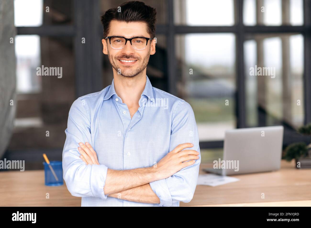 Formelles Geschäftsporträt. Der selbstbewusste, erfolgreiche kaukasische Geschäftsmann oder Manager mit Brille steht neben seinem Schreibtisch im Büro, die Arme sind gekreuzt, blickt direkt auf die Kamera und lächelt freundlich Stockfoto