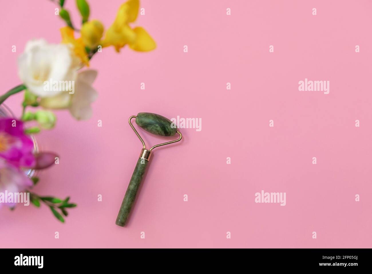 Beauty Jade Roller mit Blumen auf pastellrosa Hintergrund, Draufsicht, selektiver Fokus Stockfoto