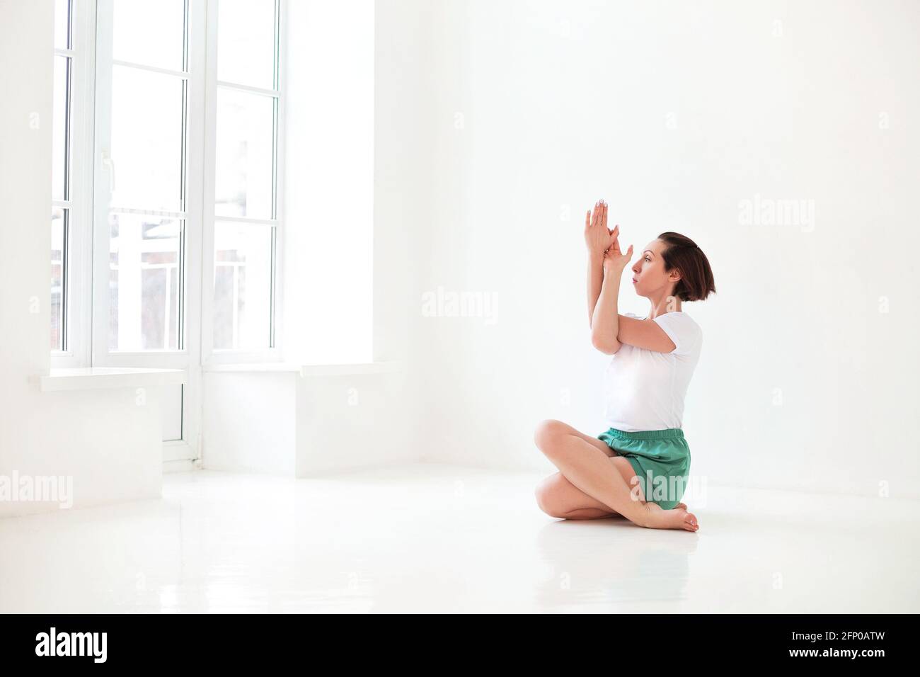 Junge Brünette weiblich in weißen Sportbekleidung auf Matte und tun Yoga beim Üben von Achtsamkeit im hellen Raum Stockfoto