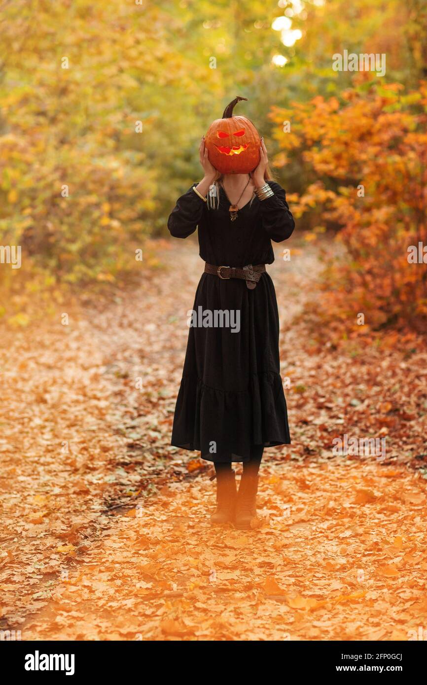 Junge Frau in schwarzem Umhang mit gruseligen Jack o Laterne Und auf den Halloween-Tag im Herbstwald wegschauen Stockfoto