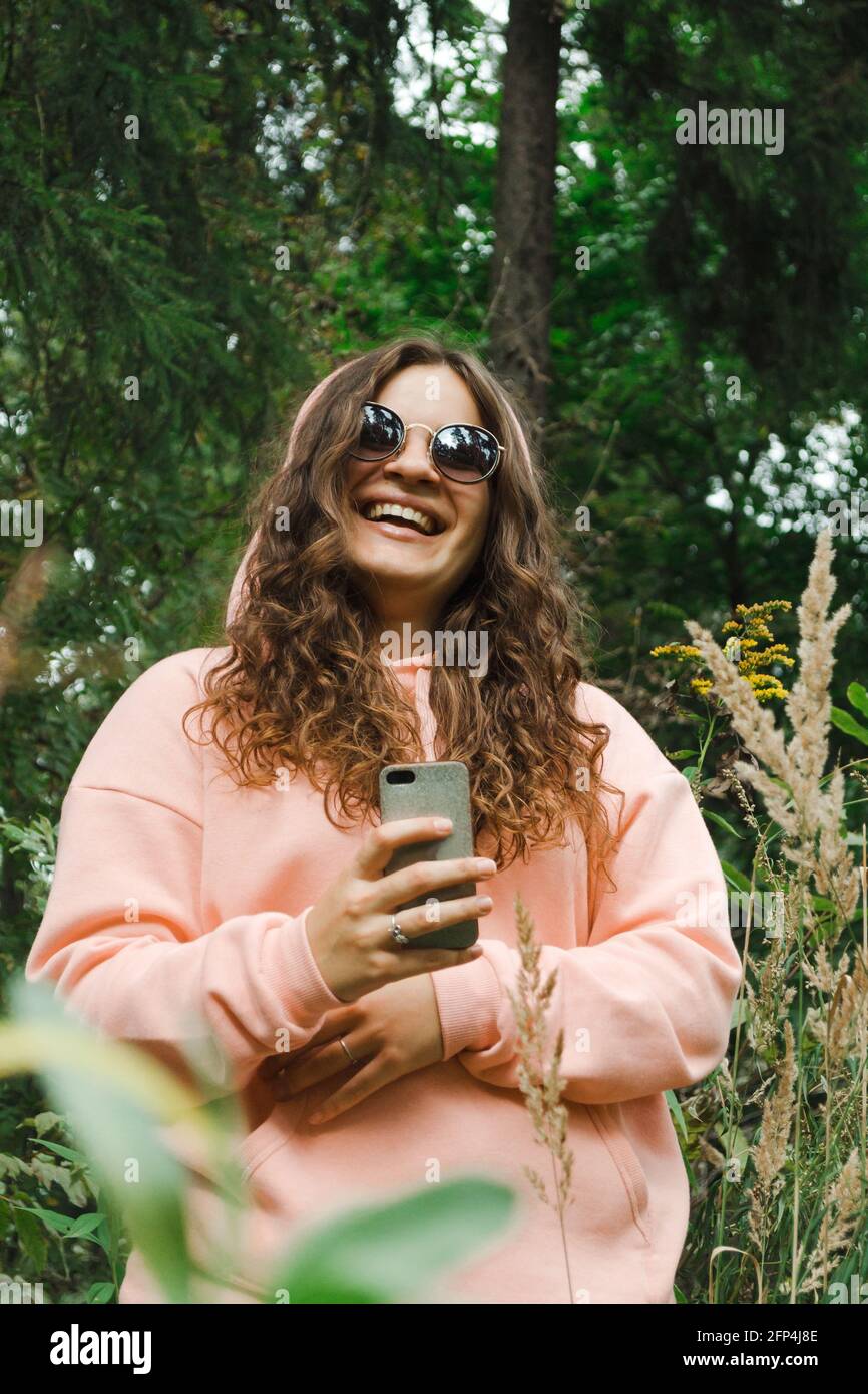 Eine junge Frau in einem rosa Sweatshirt, in einer Sonnenbrille, mit einem Telefon, lacht Stockfoto