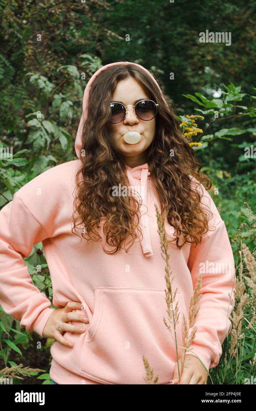 Junge Frau in einem rosa Hoodie, in einer Sonnenbrille, die eine Blase bläst. Stockfoto