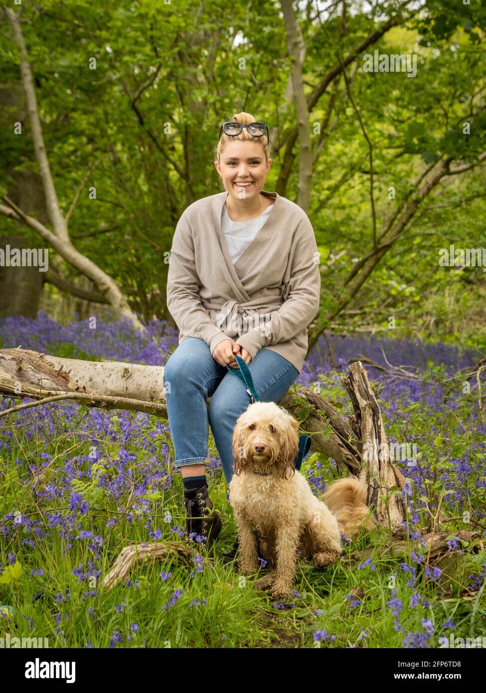 Lässig gekleidete, blonde kaukasische Hündin, die mit einem Hund auf einem Baumstamm sitzt, in einem mit Blaubellen ausgelegten Holz. Stockfoto