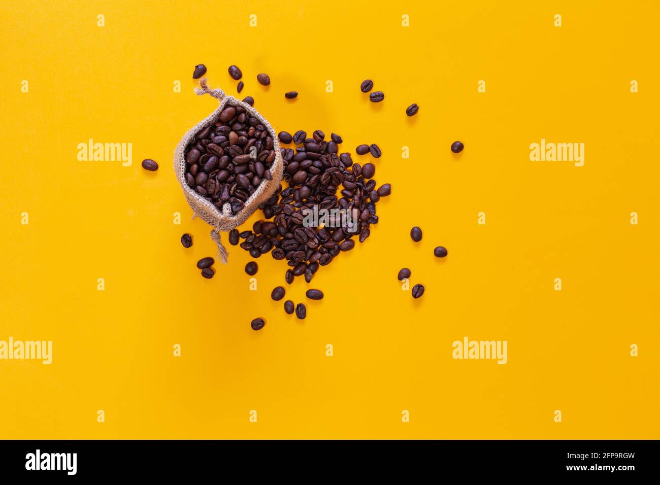 Ein Stoffsack voller Kaffeebohnen und daneben viele Bohnen, die auf gelbem Hintergrund verstreut sind. Es wird von oben betrachtet und es gibt Platz für Text oder l Stockfoto