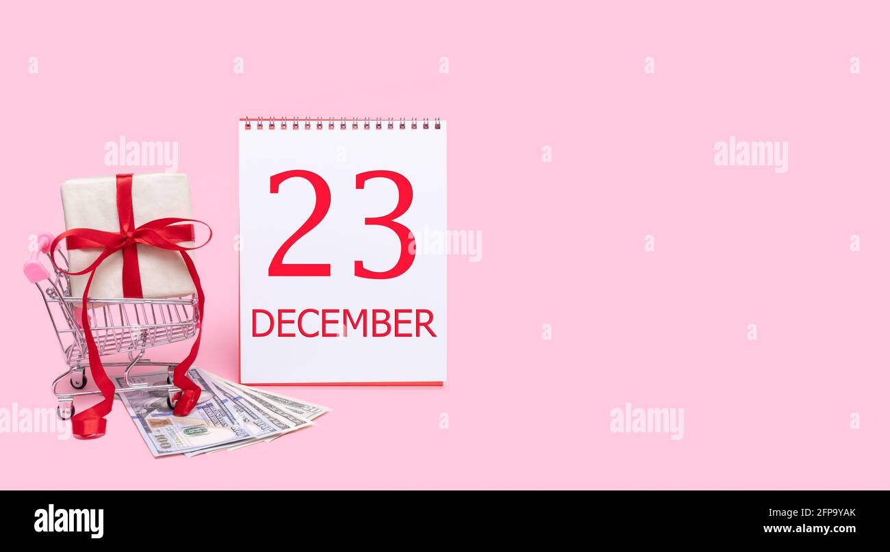 Tag des 23. dezember. Eine Geschenkbox in einem Einkaufswagen, Dollars und ein Kalender mit dem Datum des 23. dezember auf einem rosa Hintergrund. Wintermonat, Tag von Stockfoto
