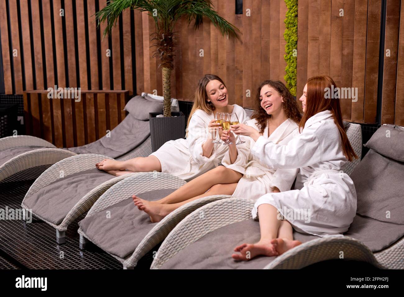 Drei Frauen in weißen Bademänteln sitzen auf einer Chaiselongue und trinken Champagner im Spa, feiern, unterhalten sich und klirren in Gläsern Stockfoto