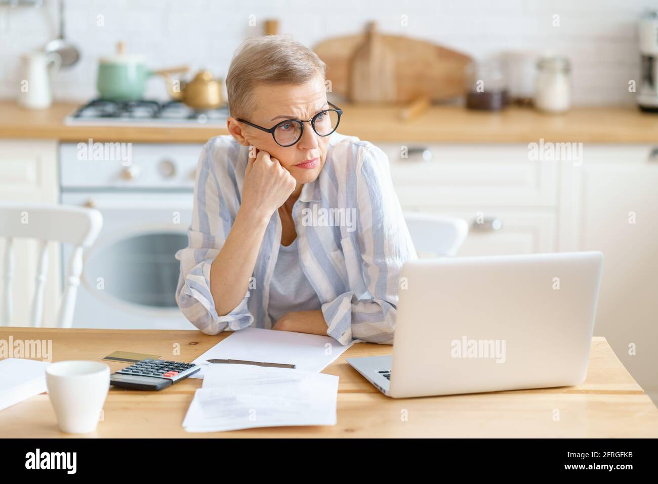 Frustrierte, reife Frau, die eine Brille trägt und Dokumente analysiert, und sich aufgrund der hohen Kosten für die Hypothekenzahlung traurig, aufgeregt und müde fühlt. Arbeiten mit einem Laptop von zu Hause aus. Senior People und freiberufliches Konzept Stockfoto