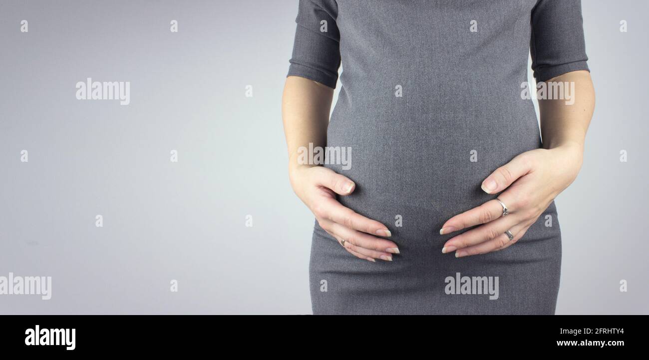 Eine Schwangere im Kleid hält die Hände auf dem Bauch auf einem grauen Hintergrund. Mutti. Stockfoto