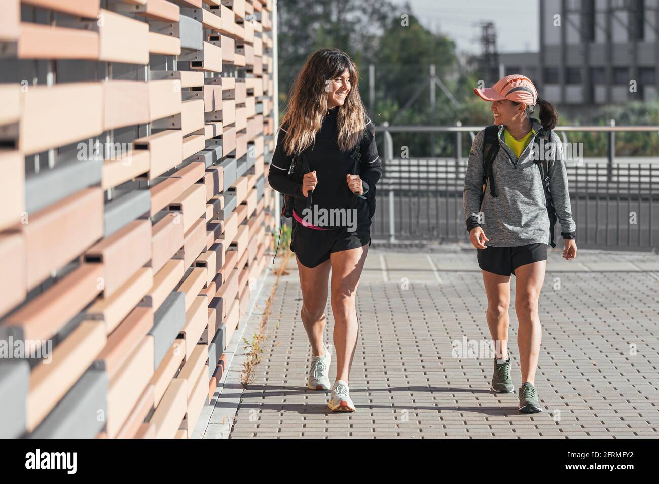 Lächelnde, multiethnische Frauen bereiten sich auf einen gemeinsamen Lauf vor. Zwei Mädchen, die nach einer Outdoor-Trainingseinheit spazierengehen Stockfoto