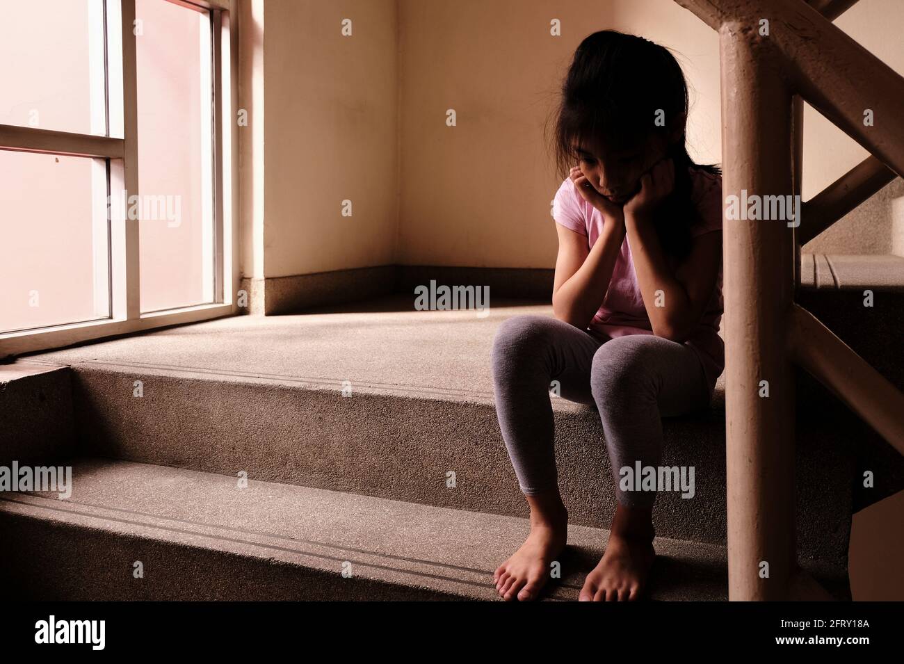 Konzept für Kindesmissbrauch. Ein trauriges und einsames junges Mädchen, das an einer Treppe in einem Gebäude sitzt, mit Licht durch ein Glasfenster im Hintergrund. Stockfoto