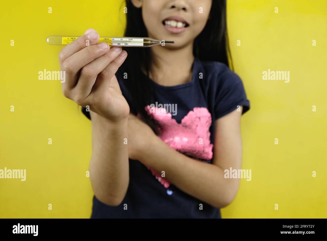 Ein junges asiatisches Mädchen hält in einer Hand ein Quecksilberthermometer mit Glasröhrchen hoch und zeigt die Temperatur an. Hellgelber Hintergrund. Stockfoto