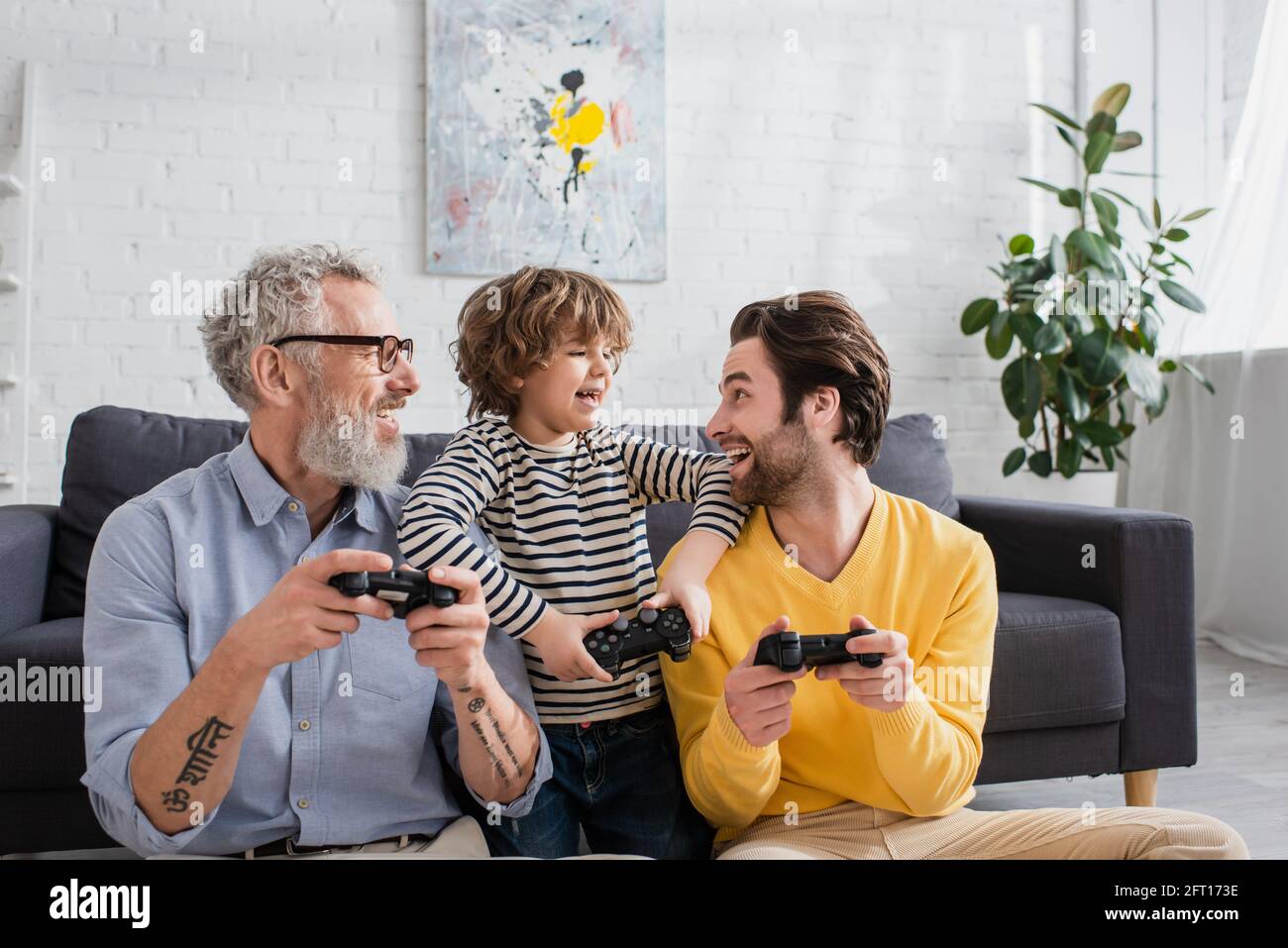 KIEW, UKRAINE - 12. APRIL 2021: Fröhliches Kind, das Vater mit Joystick in der Nähe des Großvaters anschaut Stockfoto