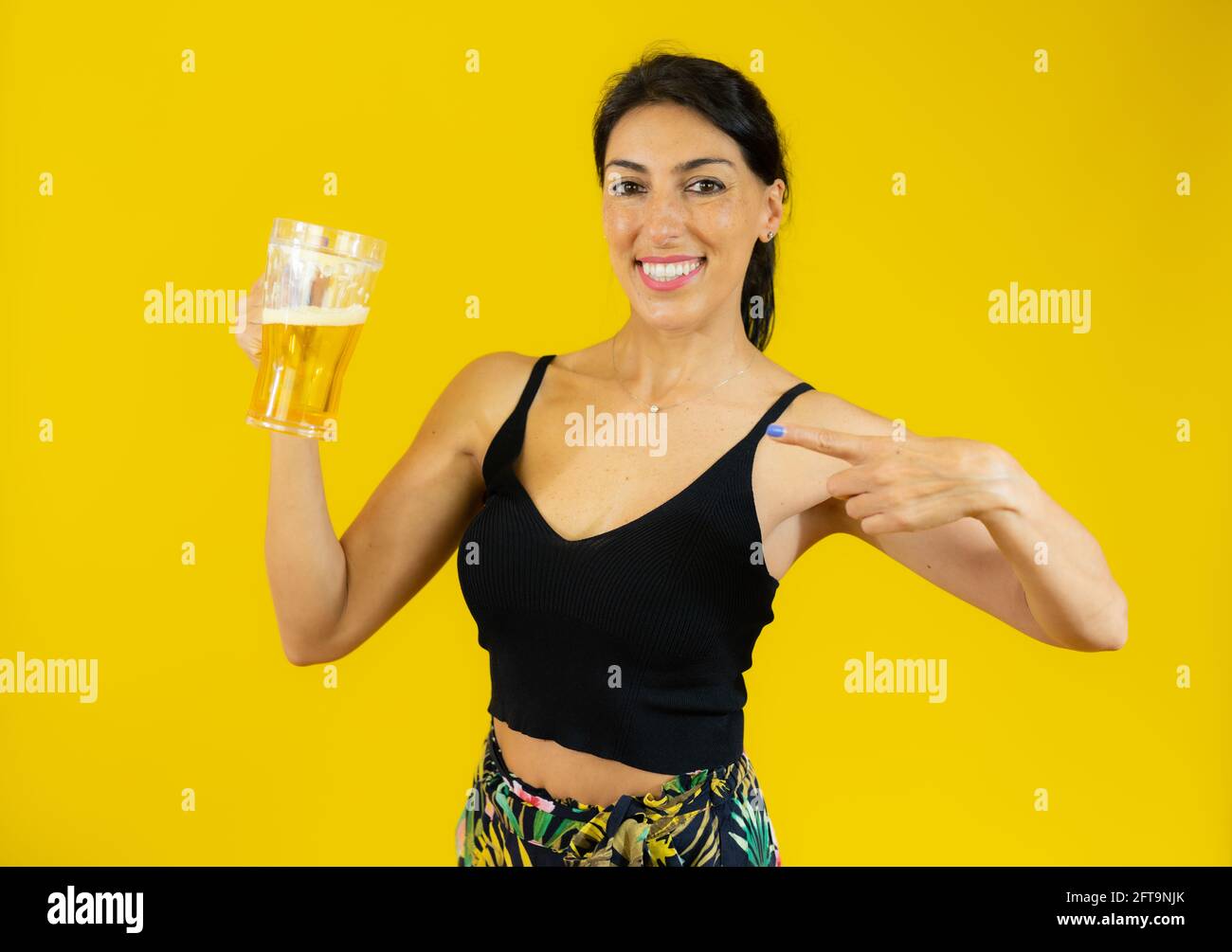 Junge Frau in Sommerkleidung trinkt Glas Bier stehend Über isoliertem gelben Hintergrund Stockfoto