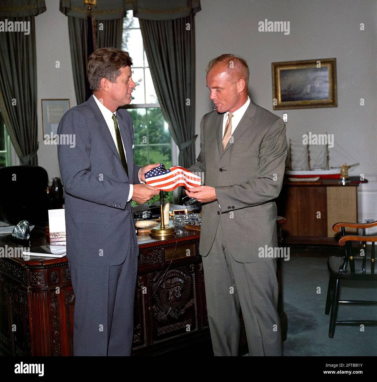 Präsident John F. Kennedy erhält vom Astronauten-Oberstleutnant John H. Glenn, Jr. (rechts) ein Geschenk einer amerikanischen Flagge; LT. Col. Glenn trug die Flagge in seinem Raumanzug während seines Orbitalfluges an Bord von Mercury-Atlas 6, auch bekannt als Friendship 7. Der Sonderassistent des Präsidenten, Kenneth P. O'Donnell, steht im Hintergrund. Oval Office, White House, Washington, D.C. Stockfoto