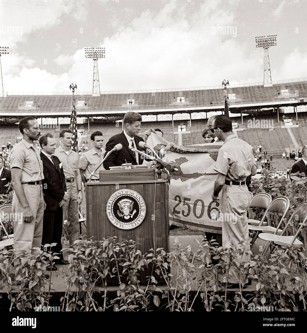 Präsident John F. Kennedy erhält die Flagge der 2506. Kubanischen Invasionsbrigade von Brigade-Mitgliedern während einer feierlichen Präsentation im Orange Bowl Stadium in Miami, Florida. Auf dem Rednerpult (von links nach rechts): Erneido Oliva, zweiter Kommandant der Brigade; Robert King High, Bürgermeister von Miami; Jose Pérez San Román, Kommandeur der Brigade; Manuel Artime, Zivilchef der Brigade; Präsident Kennedy (am Rednerpult mit Flagge); Dolmetscher des US-Außenministeriums, Donald Barnes (meist hinten versteckt, mit Flagge); First Lady Jacqueline Kennedy – teilweise hinter Flagge versteckt. Stockfoto
