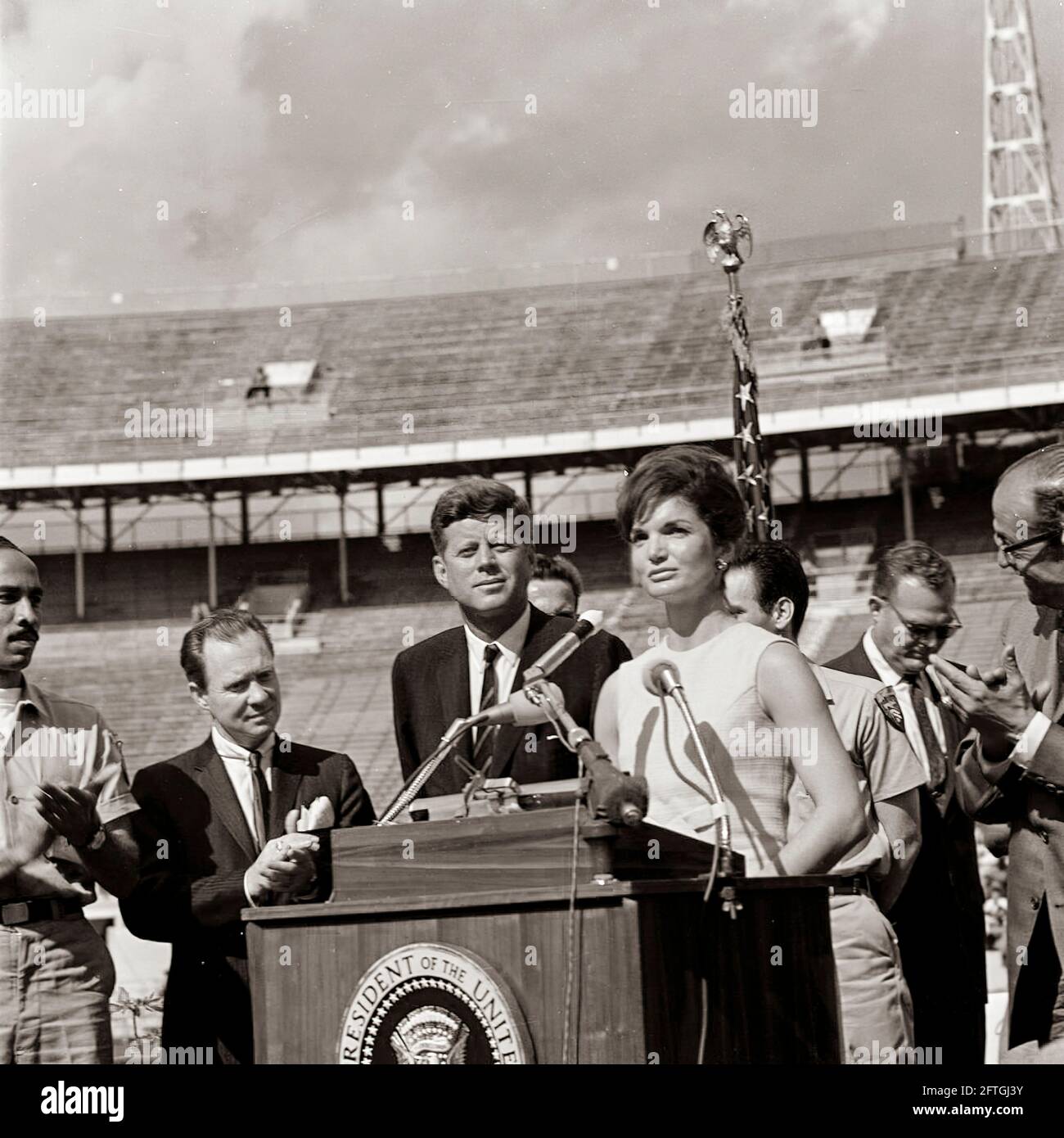 First Lady Jacqueline Kennedy hält bei einer Zeremonie der 2506. Flagge der kubanischen Invasionsbrigade im Orange Bowl Stadium in Miami, Florida, Bemerkungen auf Spanisch; Mitglieder der Brigade überreichten ihre Flagge an Präsident John F. Kennedy und Frau Kennedy. Auf der Rednerplattform (von links nach rechts): Erneido Oliva, zweiter Kommandant der Brigade (applaudierende); Bürgermeister von Miami, Robert King High (applaudierende); Präsident Kennedy; Jose Pérez San Román, Kommandant der Brigade (hinten, meist versteckt); die First Lady (an Rednermikrofonen); Manuel Artime, Chef der zivilen Brigade . Stockfoto