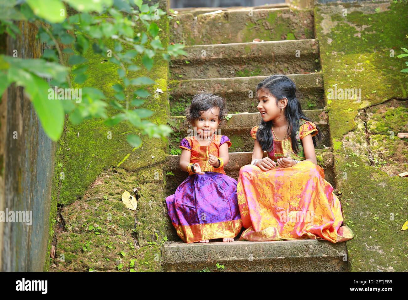 Südindische Mädchen Kinder tragen schöne traditionelle Kleid langen Rock Und Bluse, sitzend auf den langen Stufen eines bedeckten Hauses Mit moosgrünem Pilz Stockfoto