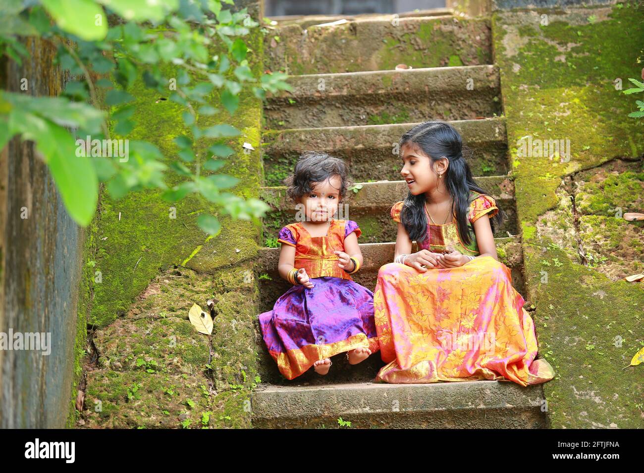 Südindische Mädchen Kinder tragen schöne traditionelle Kleid langen Rock Und Bluse, sitzend auf den langen Stufen eines bedeckten Hauses Mit moosgrünem Pilz Stockfoto