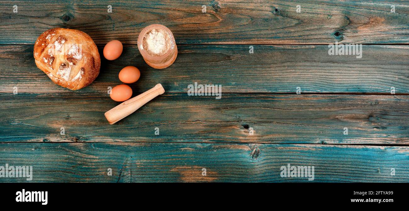 Frisch gebackener ganzer Laib Sauerteig, Mehl, Eier und Nudelholz auf verblassten blauen Holzplanken im flachen Lay-Format Stockfoto