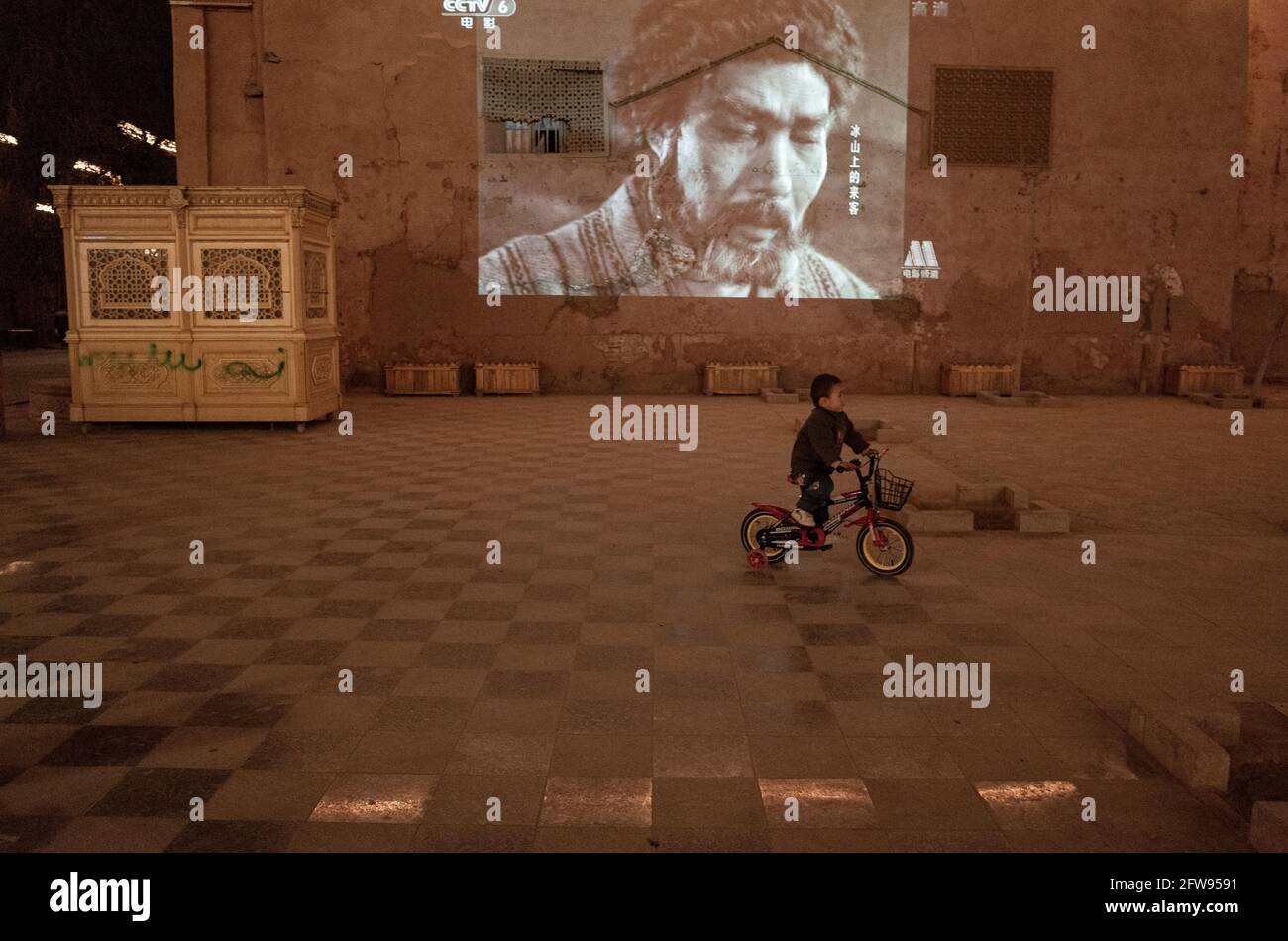 Junge spielt auf seinem Fahrrad auf dem Platz, während im Hintergrund ein Film über die chinesische Revolution gezeigt wird. Kashagar China 2019 Stockfoto