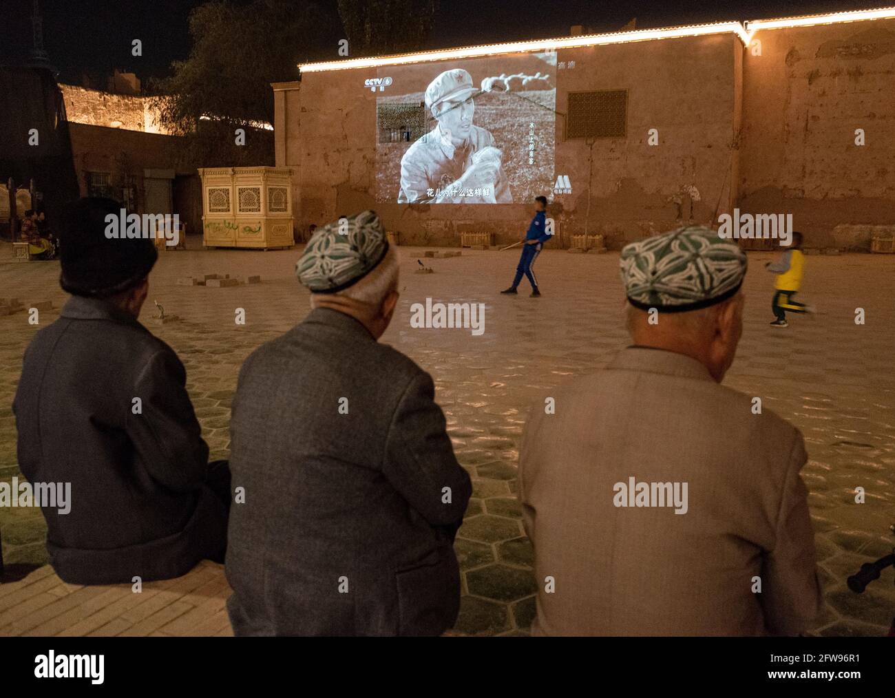 Uyhgur Männer sitzen auf einem kleinen Platz und schauen sich einen Propagandafilm über die Chinesische Revolution an. Kashgar , Volksrepublik China 2019 Stockfoto