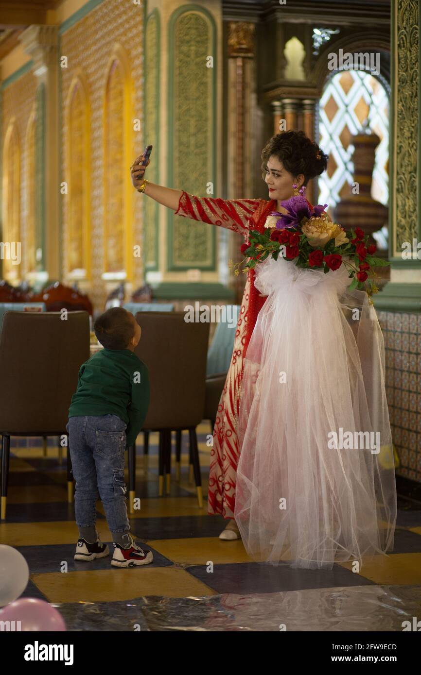 Junge, der seine Mutter ansieht, die ein Foto mit ihrem Telefon macht Kashgar, Xinkiang, Volksrepublik China, 2019 Stockfoto