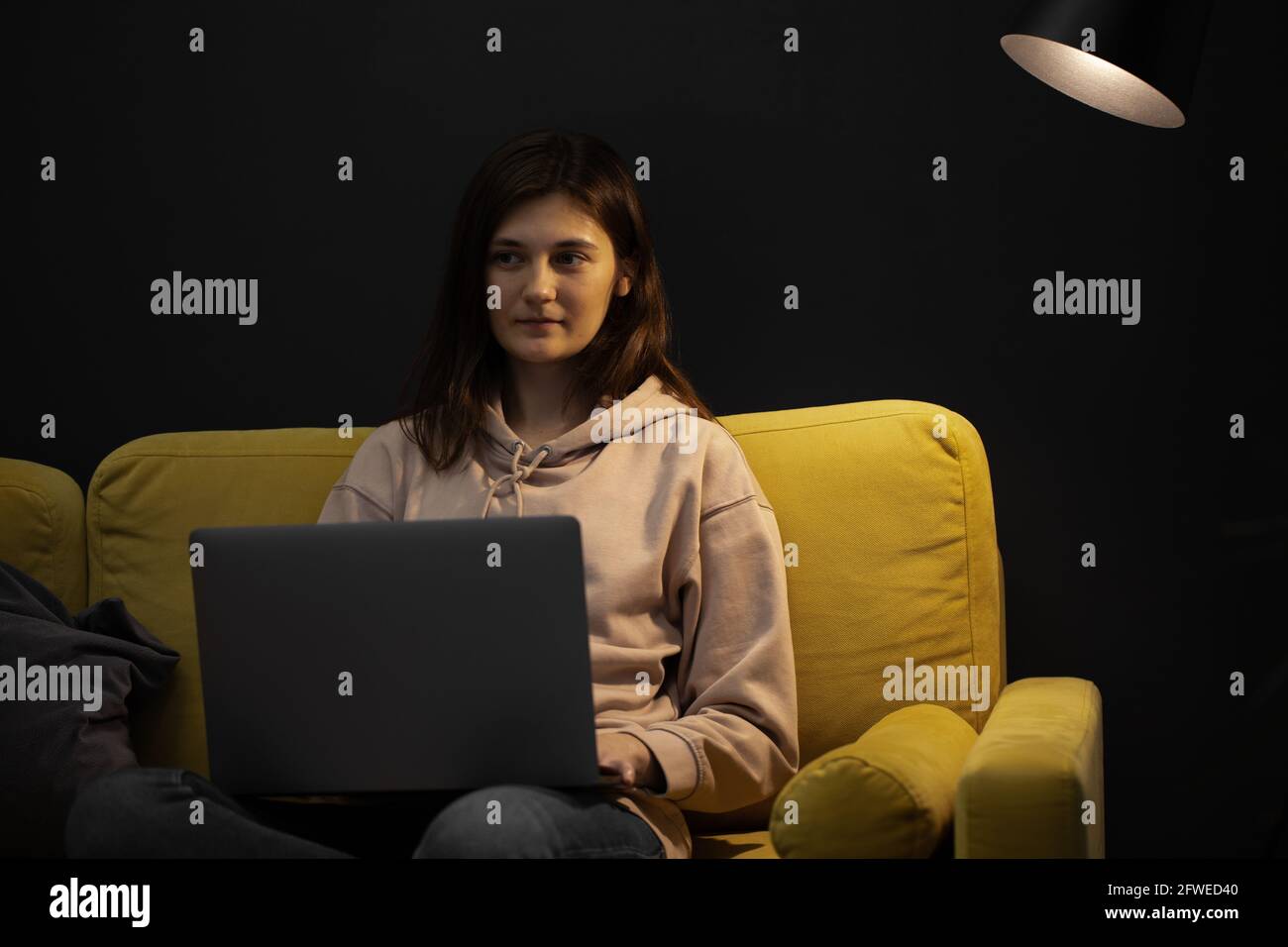 Langhaariges Mädchen sitzt nachdenklich auf der Couch mit Laptop unter Punkt Lampe Stockfoto