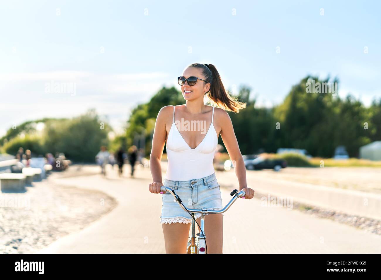 Hübsche Frau, die Fahrrad im Stadtpark Radweg und Straße reitet. Fahrradpromenade im Sommer. Fröhlicher Radfahrer. Modische, trendige Jeans-Shorts. Stockfoto