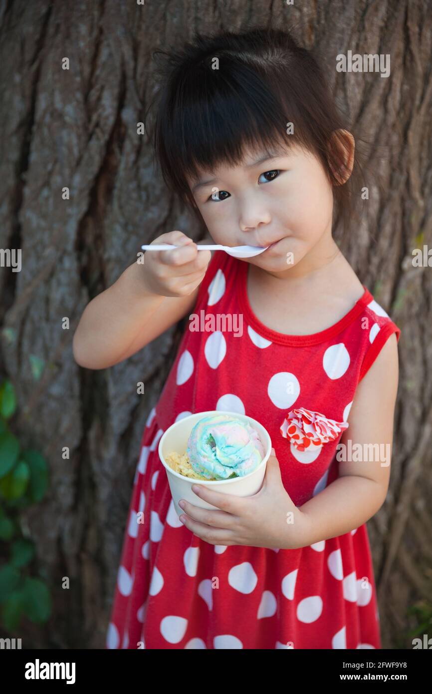 Liebenswert asiatische Mädchen essen Eis im Sommer im Urlaub über einem Stamm eines Baumes Hintergrund. Hübsches Kind genießen und entspannen in der Natur. Im Freien. Stockfoto