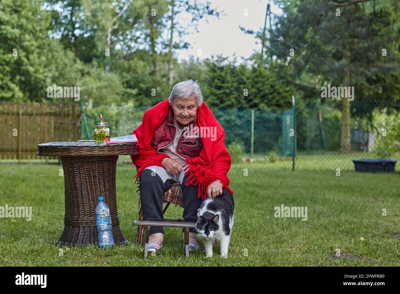Ältere Frau, die draußen auf dem Pensionat sitzt, in rotem Poncho mit schwarz-weißem Katzenhüter Stockfoto