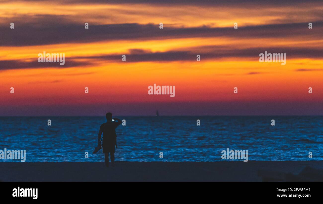 Der Mann fotografiert mit dem Telefon im Urlaub am Strand in Silhouette am Strand gegen den lebhaften Sonnenuntergang am Himmel in der Abenddämmerung. Stockfoto