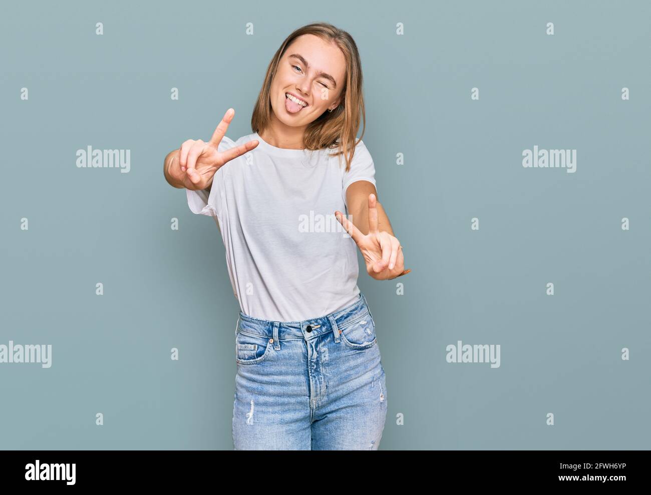 Schöne junge blonde Frau trägt beiläufige weiße T-Shirt lächelnd mit Zunge  aus zeigt Finger von beiden Händen tun Siegeszeichen. Nummer zwei  Stockfotografie - Alamy
