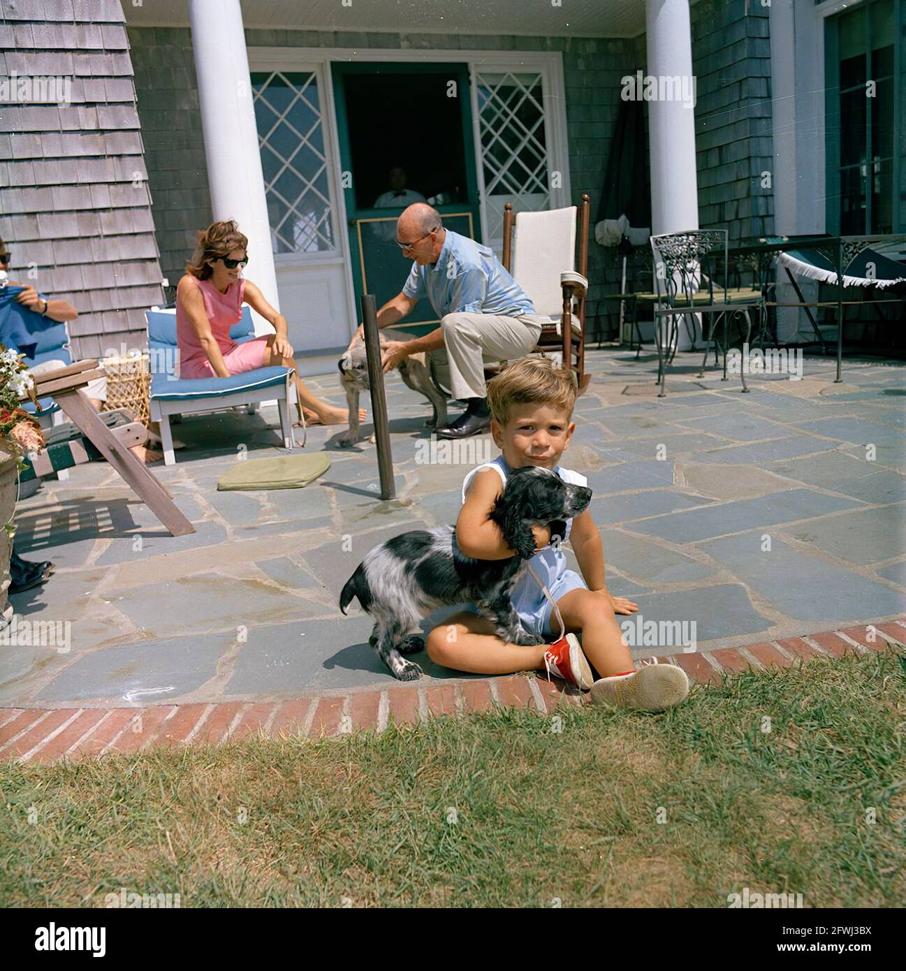 John F. Kennedy, Jr., spielt mit seinem Hund Shannon im Brambletyde-Haus auf Squaw Island, Hyannis Port, Massachusetts. Präsident John F. Kennedy telefoniert am linken Rand des Rahmens; First Lady Jacqueline Kennedy und der Assistentin des Präsidenten, Dave Powers, spielen im Hintergrund mit dem Hund der Kennedy-Familie, Wolf. Stockfoto