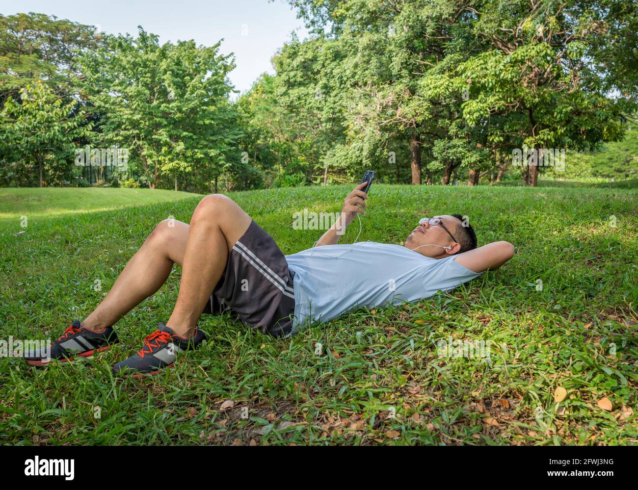 Der asiatische Jogger mittleren Alters legte sich in einem öffentlichen Park auf den Rasen und benutzte ein Mobiltelefon mit Kopfhörer. Stockfoto