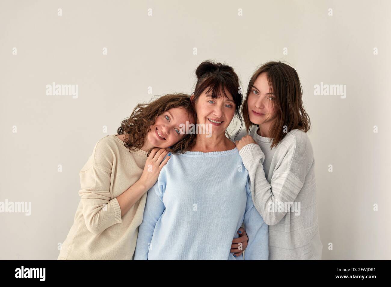 Fröhliche Erwachsene Mutter umarmt charmante junge Töchter im Stehen Weißer Hintergrund im Studio und Blick auf die Kamera Stockfoto
