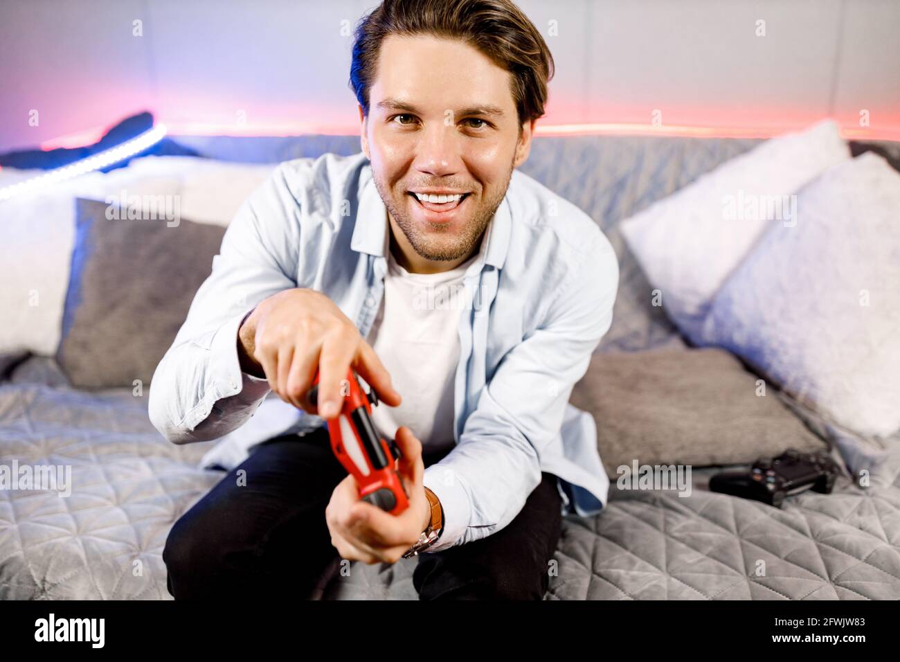 Nahaufnahme attraktiver junger Mann, hält einen Joy Stick und spielt Videospiele im Fernsehen, sitzt zu Hause auf einem gemütlichen Sofa Neon, erfolgreich, gewinnt er ein verrücktes Auto Stockfoto Nahaufnahme attraktiver junger Mann, hält einen Joy Stick und spielt Videospiele im Fernsehen, sitzt zu Hause auf einem gemütlichen Sofa Neon, erfolgreich, gewinnt er ein verrücktes Auto Stockfoto