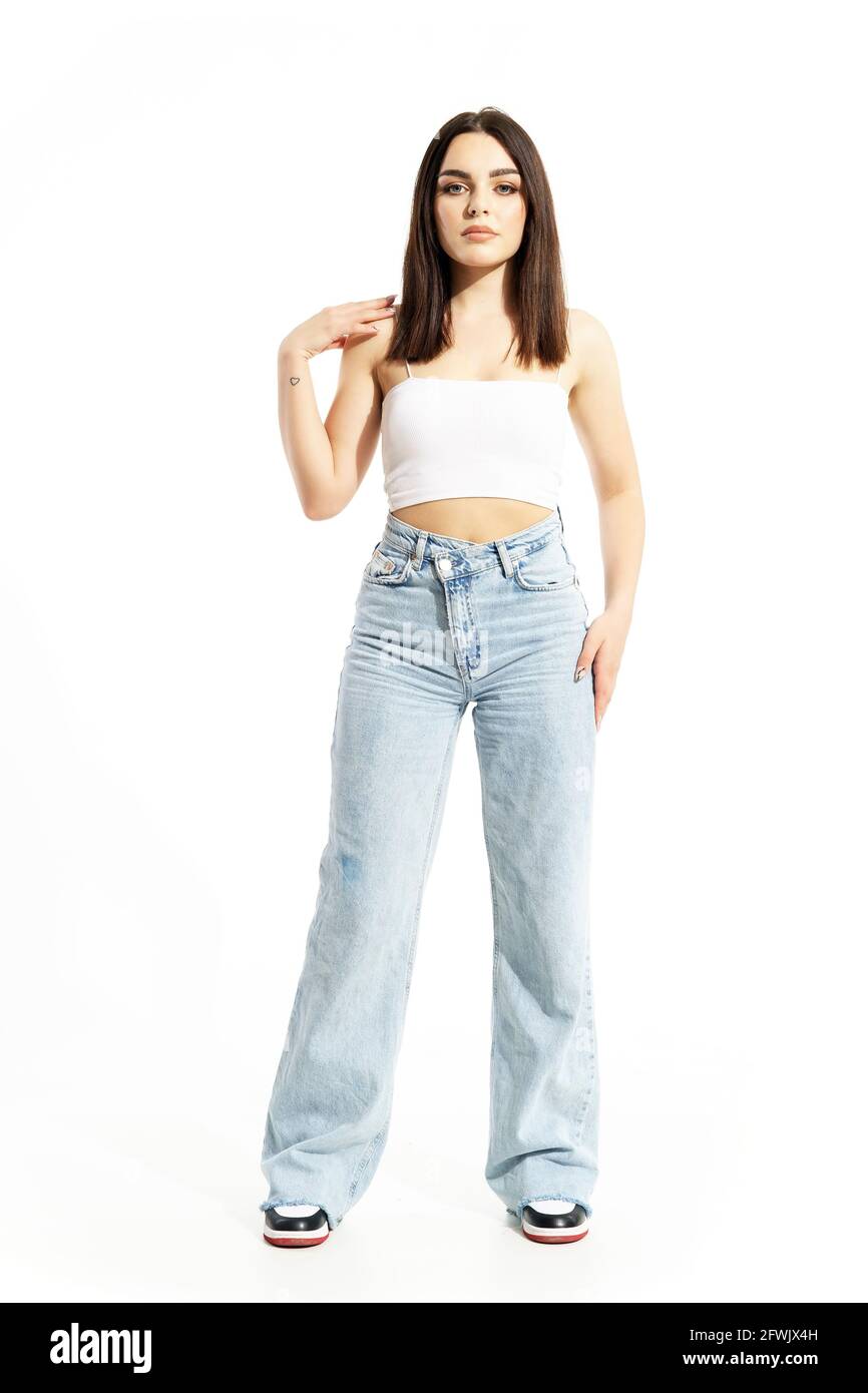 Gen Z feminine junge selbstbewusste Frau posiert berührende Schulter in weißem Oberteil und Jeans. Ganzkörperlänge isoliert auf weißem Hintergrund Stockfoto