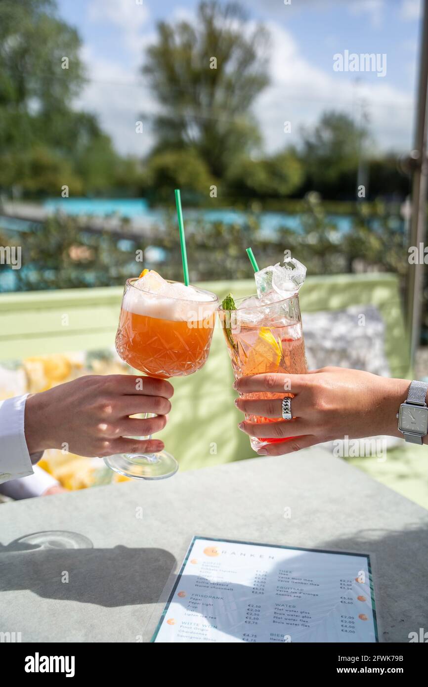 Zwei Personen jubeln mit Cocktails draußen auf einer sonnigen Terrasse und einem blauen Pool im Hintergrund Stockfoto