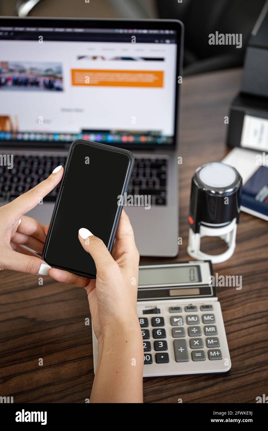 Smartphone in Frauenhand am Schreibtisch vor dem Hintergrund eines Rechners und eines Laptops. Stockfoto