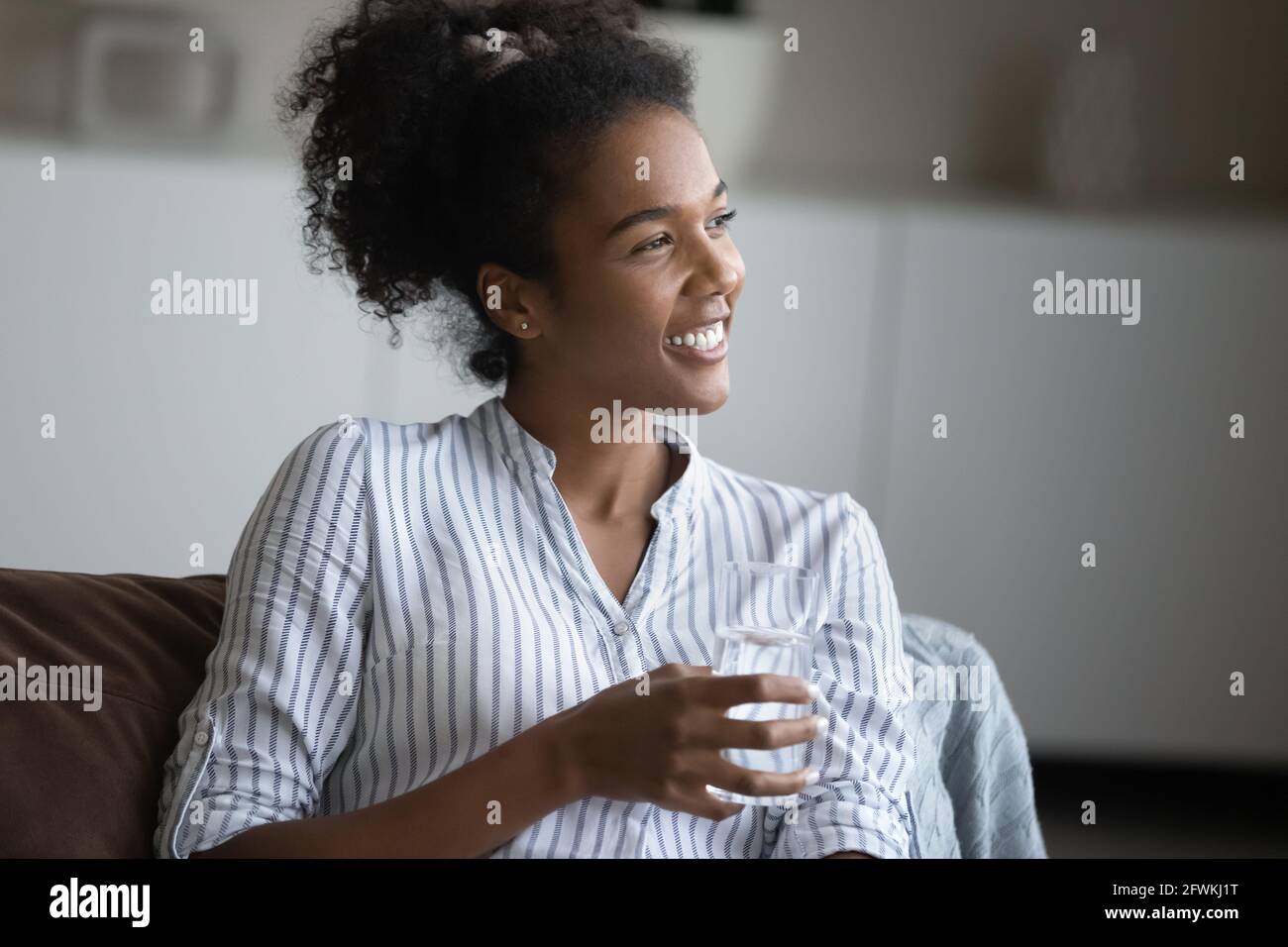 Lächelnde, verträumte junge afrikanische Frau mit einem Glas Mineralwasser. Stockfoto