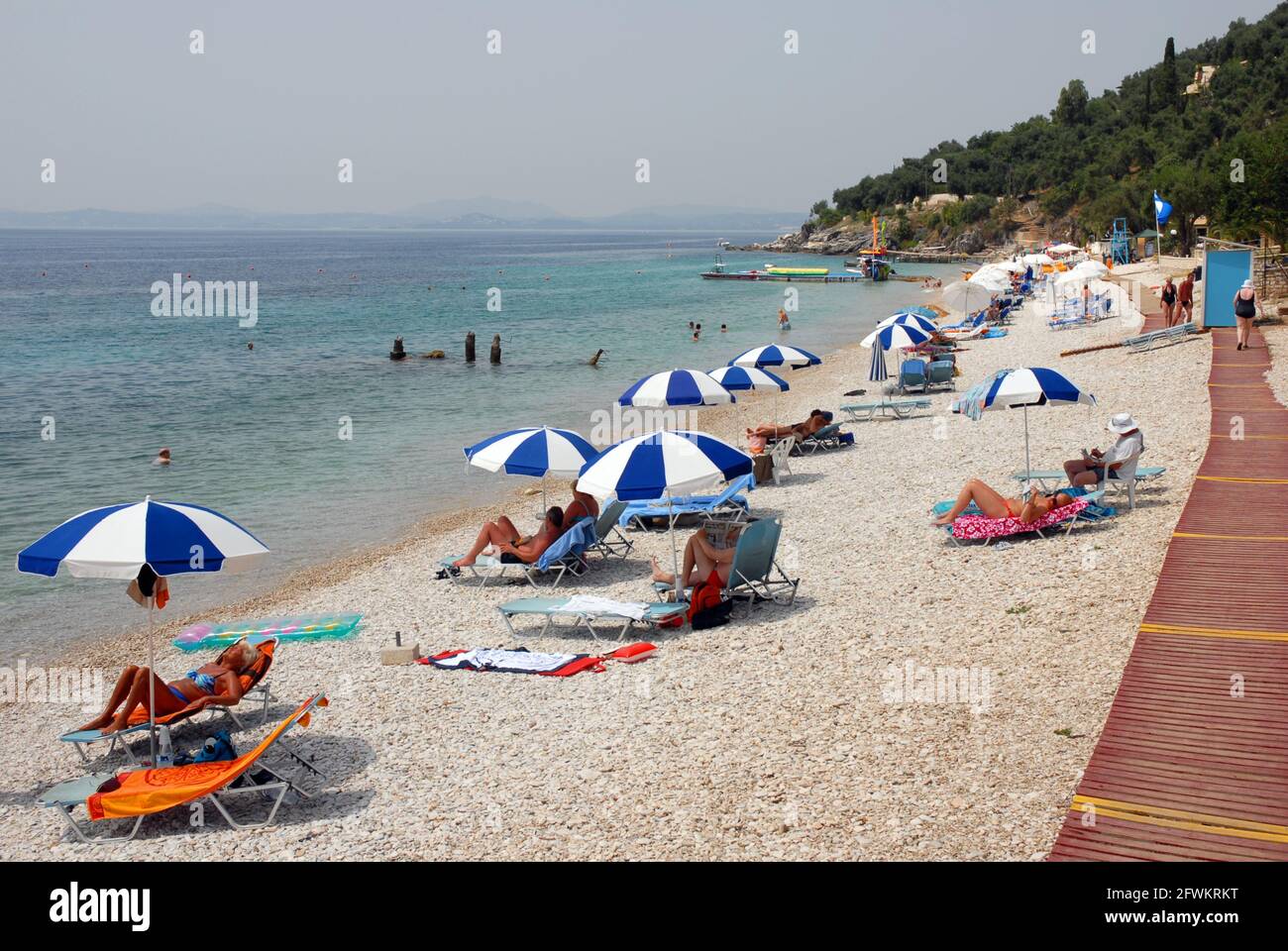 Leute zur freien Verfügung, genießen die heiße Sonne, Nissaki Beach, der mit zweckbestimmten hölzernen Gehwegen über dem Schindel versehen wird, Korfu, Griechenland Stockfoto
