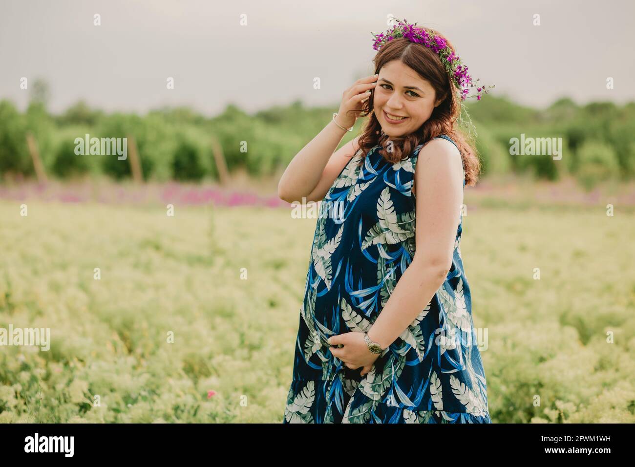 Nahaufnahme einer jungen lächelnden Schwangeren mit wilden Blumen Auf dem Kopf genießen Mutterschaft im Feld stehen und betrachten Kamera Stockfoto