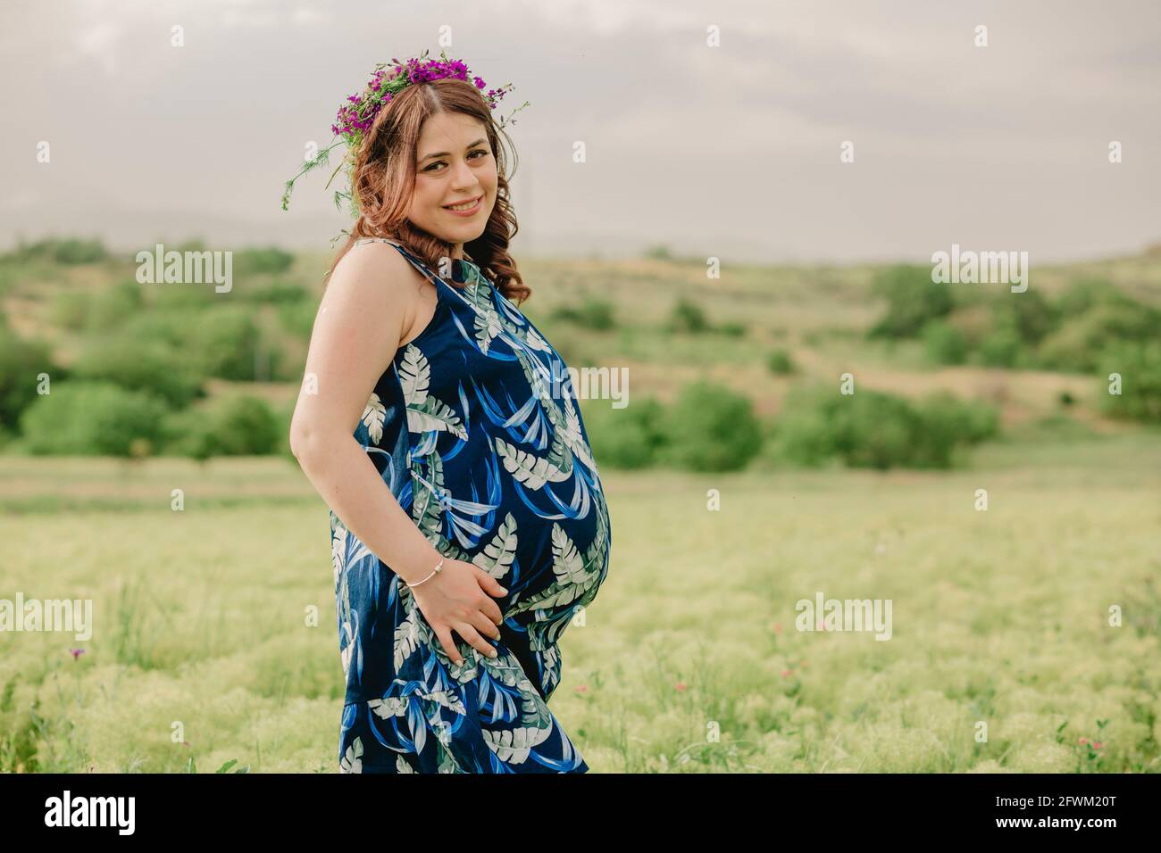 Schwangerschaft und Mutterschaft. Junge, schwanger Frau mit wilden Blumen auf dem Kopf, die auf dem Feld stehen, ihren großen Bauch halten und für die Kamera posieren Stockfoto