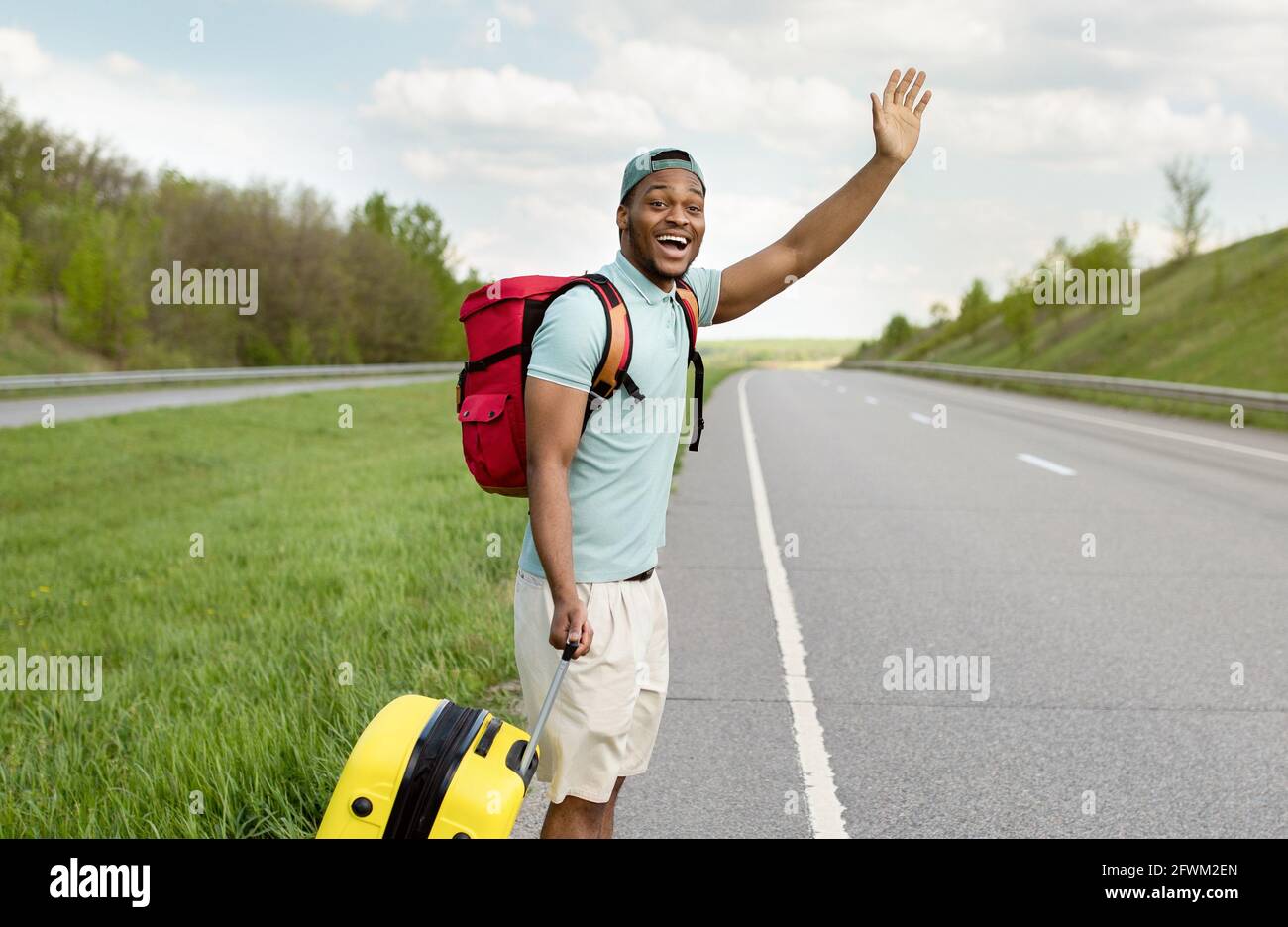 Attraktiver schwarzer Mann mit Gepäck winkt ein Auto herunter, fährt am Straßenrand mit Anhalter, fährt mit Autostop, im Freien Stockfoto