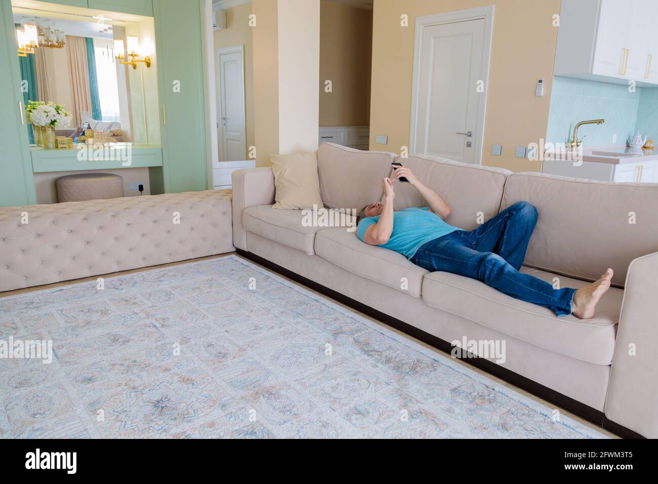 Ein erwachsener Mann durchsucht in einem Mobiltelefon soziale Netzwerke, während er auf einem Sofa in einer Wohnung liegt. Stockfoto
