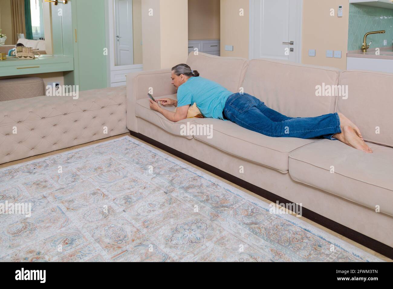 Ein erwachsener Mann durchsucht in einem Mobiltelefon soziale Netzwerke, während er auf einem Sofa in einer Wohnung liegt. Stockfoto