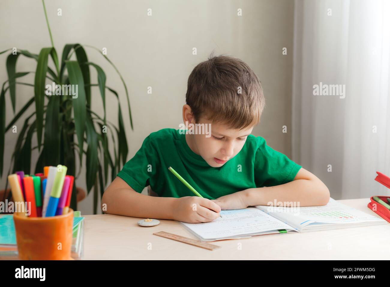 7 Jahre alter Junge, der Unterricht macht und am Schreibtisch in seinem Zimmer sitzt. Kind schreibt Hausaufgaben im Notizbuch Stockfoto
