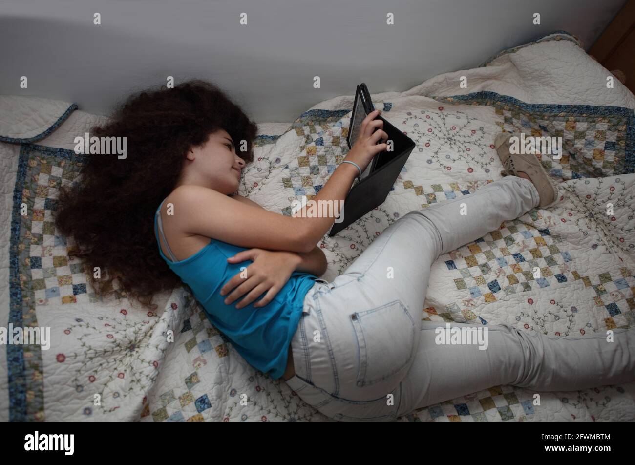 Das wunderschöne dreizehnjährige lächelnde Mädchen, Teenager mit lockigem Haar, träumt davon, auf ihrem Bett zu liegen, auf Kleidung und Schuhen, sie schaut auf ihr Tablet. Stockfoto
