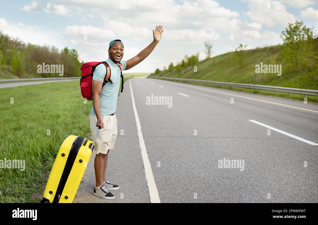 Glücklicher schwarzer Kerl mit Rucksack und Koffer, der das Auto heruntergetrampelt hat, eine Fahrt benötigt, am Straßenrand mitgetrampelt, Platz kopiert Stockfoto